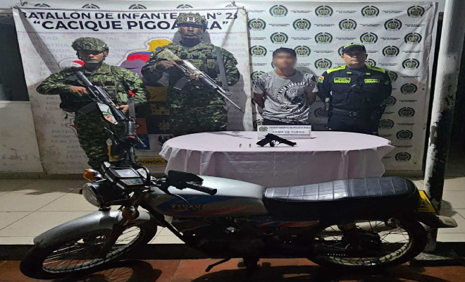 Dos funcionario del ejército, funcionario de la policía nacional y en a mitad un hombre capturado, al frente una mesa con un arma de fuego y una motocicleta color negro