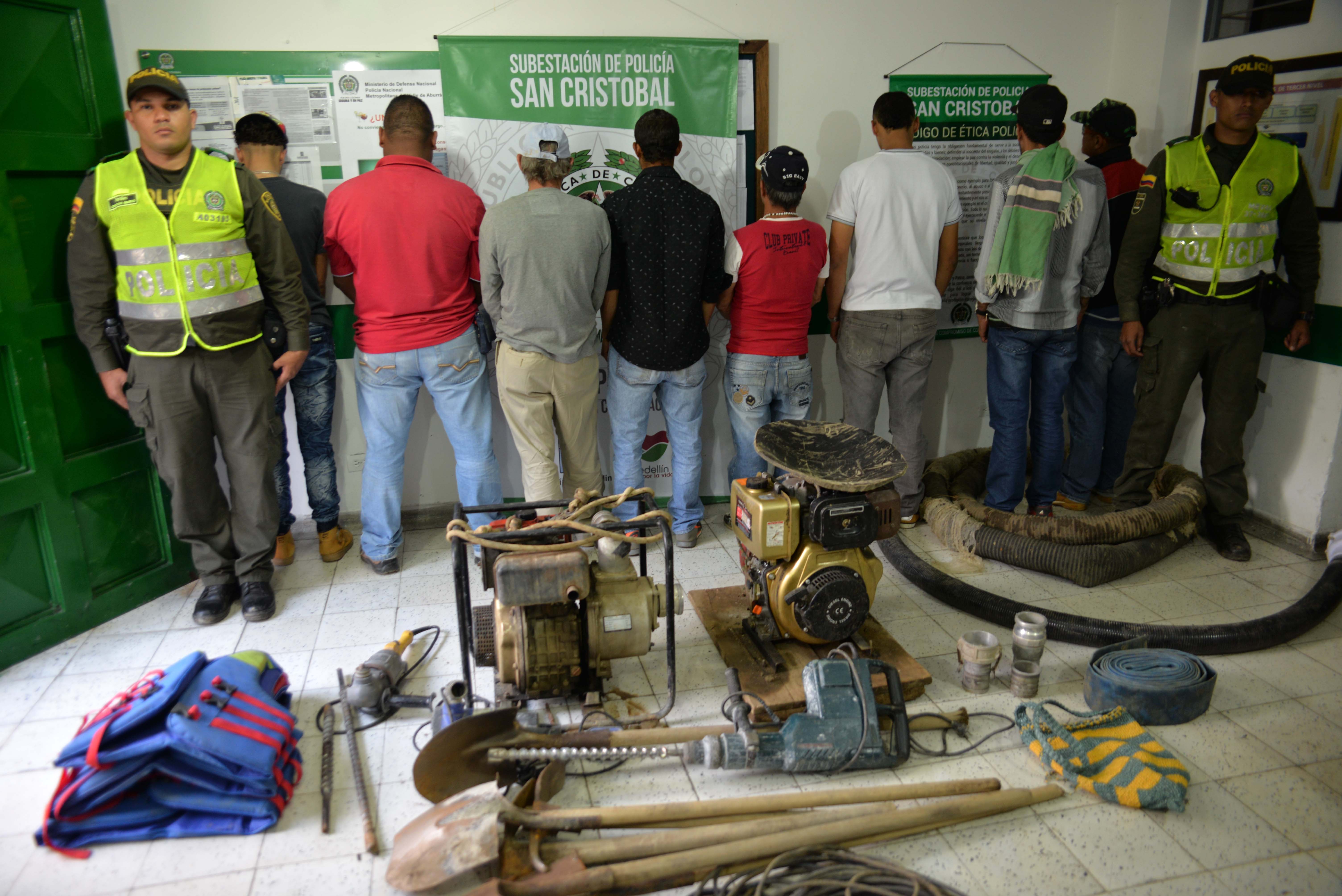 Dos policías custodian a ocho personas que están de espaldas, frente a ellos hay herramientas